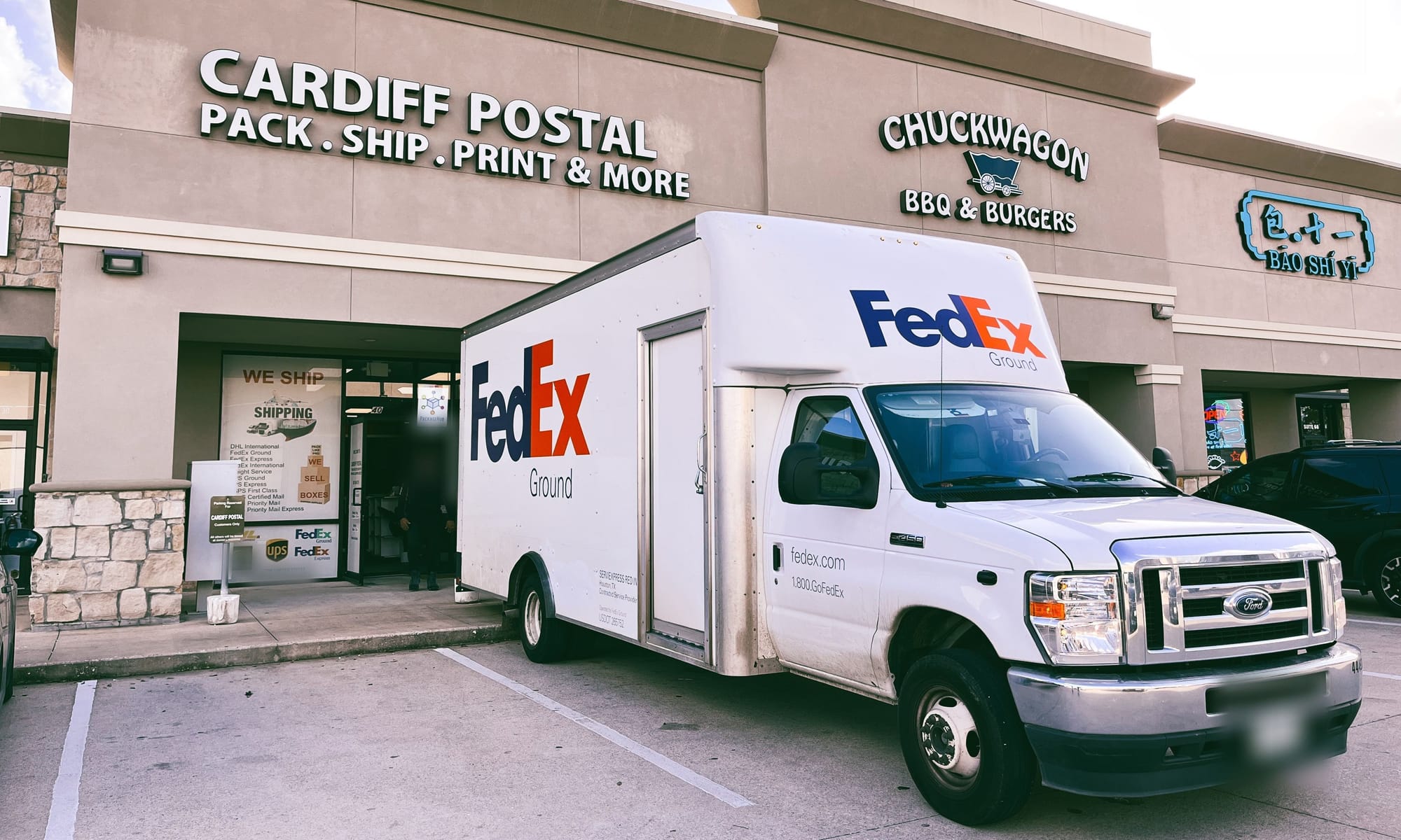A FedEx Ground box truck parked in front of Cardiff Postal, the store sign above the entrance
